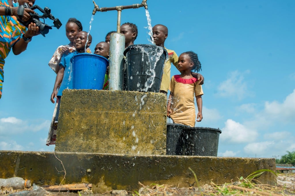 Children collecting water from a communal standpipe into buckets under a bright sky, representing community water supply.